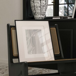 K&ouml;nnte beinhalten: Ein schwarzer gerahmter Druck mit wei&szlig;em Hintergrund und einer schwarzen und wei&szlig;en abstrakten Linienzeichnung. Der Druck lehnt an einem schwarzen Stuhl mit einer geflochtenen R&uuml;ckenlehne.