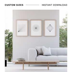 Puede incluir: Tres impresiones geométricas minimalistas en blanco y negro en marcos de madera que cuelgan de una pared blanca sobre un sofá blanco con un cojín de tiro gris claro. El sofá está frente a una ventana grande con árboles verdes afuera. Una mesa de café de madera se encuentra frente al sofá.