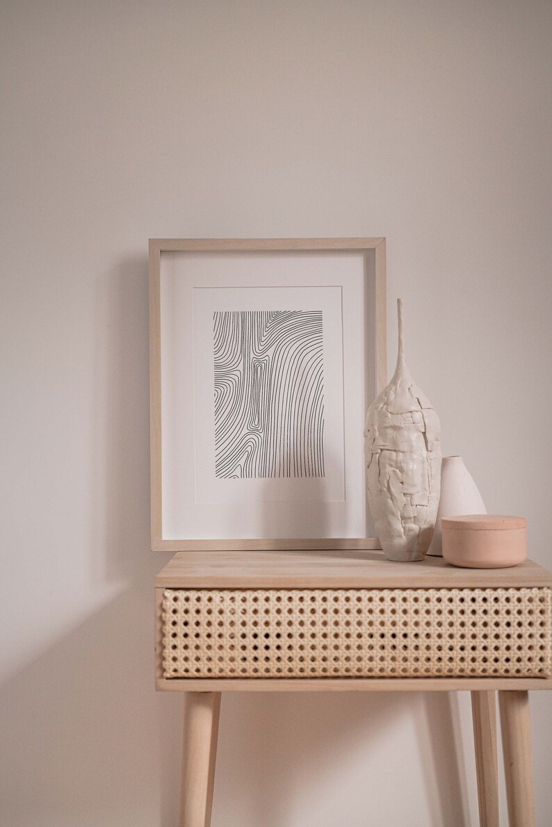 K&ouml;nnte beinhalten: Ein Holz-Beistelltisch mit einer Schublade aus geflochtenem Rattan, einem gerahmten Schwarz-Wei&szlig;-Linienkunst-Druck, einer wei&szlig;en Keramikvase, einer wei&szlig;en Keramikvase und einer hellbraunen Keramikdose.