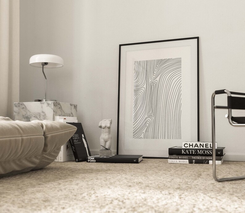 K&ouml;nnte beinhalten: Ein Schwarz-Wei&szlig;-Druck mit abstrakter Linienkunst in einem schwarzen Rahmen, der an einer wei&szlig;en Wand h&auml;ngt. Der Druck ist von B&uuml;chern und einer wei&szlig;en Lampe auf einem Marmortisch umgeben.