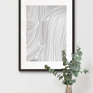 K&ouml;nnte beinhalten: Schwarz-wei&szlig;es abstraktes Linienkunst-Druckbild in einem schwarzen Rahmen, das an einer wei&szlig;en Wand h&auml;ngt. Eine Holzvase mit gr&uuml;nen Bl&auml;ttern steht auf einer wei&szlig;en Oberfl&auml;che.