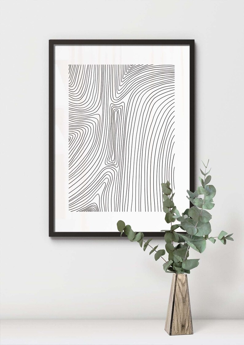 K&ouml;nnte beinhalten: Schwarz-wei&szlig;es abstraktes Linienkunst-Druckbild in einem schwarzen Rahmen, das an einer wei&szlig;en Wand h&auml;ngt. Eine Holzvase mit gr&uuml;nen Bl&auml;ttern steht auf einer wei&szlig;en Oberfl&auml;che.