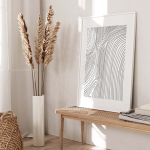 K&ouml;nnte beinhalten: Ein wei&szlig;er gerahmter Druck mit einem abstrakten Linienmuster in Schwarzwei&szlig; h&auml;ngt an einer wei&szlig;en Wand. Eine hohe Vase mit getrocknetem Pampasgras steht auf einem Holzboden neben einer hellen Holzbank.