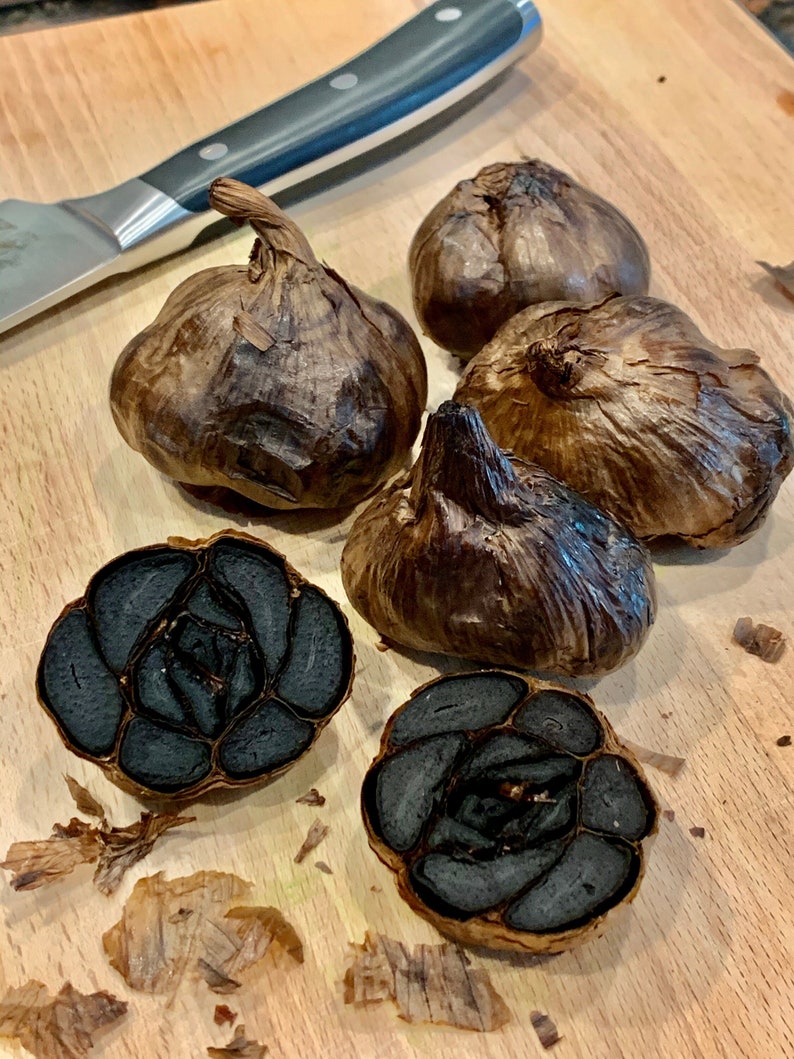 Puede incluir: Varios bulbos de ajo negro, con un cuchillo en el fondo. Un bulbo est&aacute; cortado por la mitad, revelando los dientes oscuros en el interior. El ajo tiene un color marr&oacute;n oscuro y una textura arrugada. Ingrediente culinario.