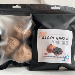 Puede incluir: Dos bolsas negras de ajo negro. Una bolsa es transparente, mostrando varios bulbos enteros. La otra bolsa tiene una etiqueta que dice "Black Garlic Whole Bulbs" con un gr&aacute;fico de un bulbo de ajo. La etiqueta tambi&eacute;n indica "Jumbo" y "Grown in American Farm, Made in America by J".