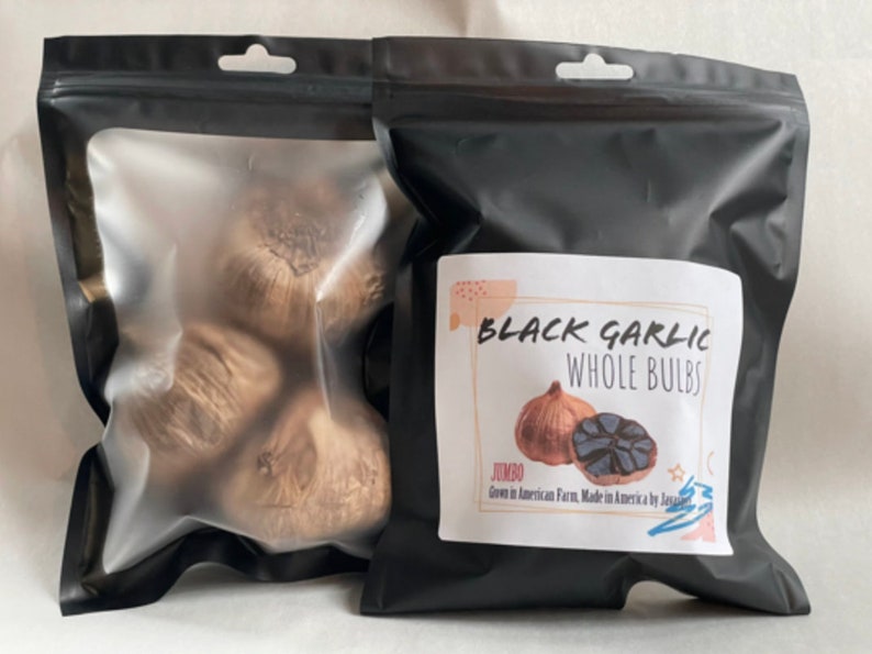 Puede incluir: Dos bolsas negras de ajo negro. Una bolsa es transparente, mostrando varios bulbos enteros. La otra bolsa tiene una etiqueta que dice "Black Garlic Whole Bulbs" con un gr&aacute;fico de un bulbo de ajo. La etiqueta tambi&eacute;n indica "Jumbo" y "Grown in American Farm, Made in America by J".