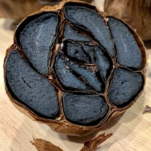 Puede incluir: Un bulbo de ajo negro cortado por la mitad, mostrando los dientes oscuros, casi negros en su interior. La piel exterior es de color marr&oacute;n claro, contrastando con el color profundo del ajo. La imagen resalta la textura y la apariencia &uacute;nica de este ingrediente especial.