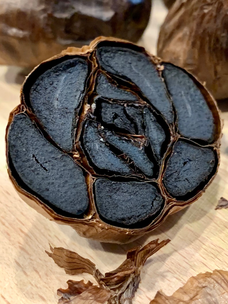 Puede incluir: Un bulbo de ajo negro cortado por la mitad, mostrando los dientes oscuros, casi negros en su interior. La piel exterior es de color marr&oacute;n claro, contrastando con el color profundo del ajo. La imagen resalta la textura y la apariencia &uacute;nica de este ingrediente especial.