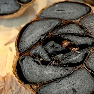 Puede incluir: Primer plano de un bulbo de ajo negro cortado por la mitad, mostrando los dientes oscuros, casi negros. Los dientes tienen una textura suave y esponjosa. La piel exterior es de color marr&oacute;n claro, contrastando con el interior oscuro.