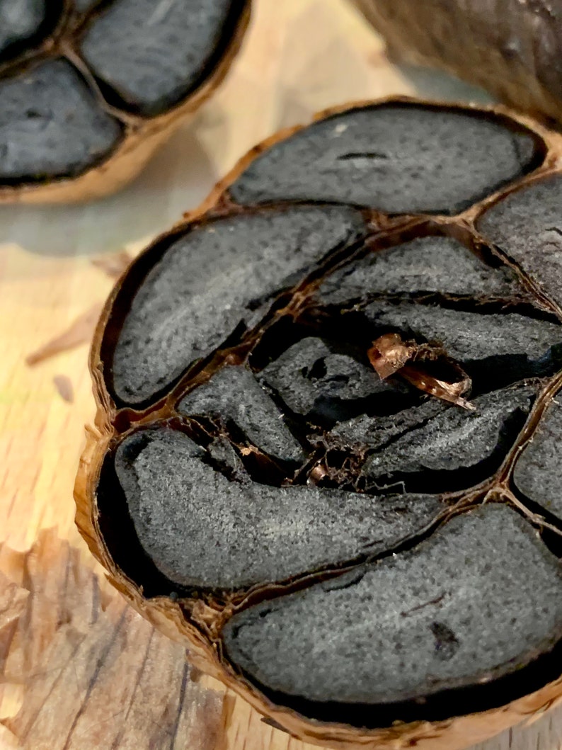 Puede incluir: Primer plano de un bulbo de ajo negro cortado por la mitad, mostrando los dientes oscuros, casi negros. Los dientes tienen una textura suave y esponjosa. La piel exterior es de color marr&oacute;n claro, contrastando con el interior oscuro.