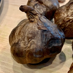Puede incluir: Primer plano de varios bulbos de ajo negro, con un exterior marr&oacute;n oscuro y arrugado. Los bulbos de ajo est&aacute;n sobre una superficie de madera de color marr&oacute;n claro. La parte superior de un bulbo tiene un tallo seco y retorcido.