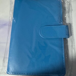 May include: A bright blue, rectangular notebook or tablet cover with a snap closure. The cover is made of a smooth material and has white stitching around the edges. The item is packaged in clear plastic.