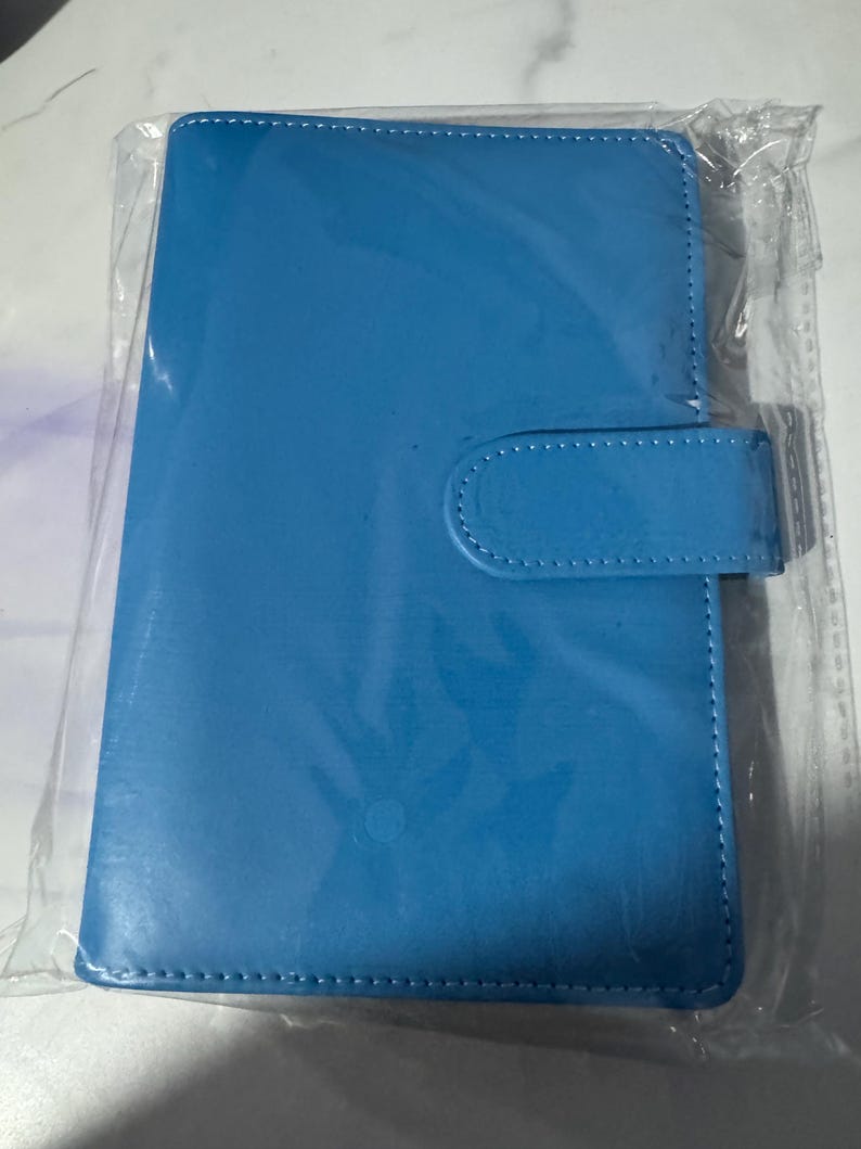 May include: A bright blue, rectangular notebook or tablet cover with a snap closure. The cover is made of a smooth material and has white stitching around the edges. The item is packaged in clear plastic.