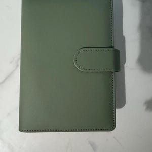 May include: A sage green, rectangular notebook with a rounded flap closure. The cover is smooth and the edges are stitched with a contrasting thread. The notebook is closed with a matching flap and a rounded tab.