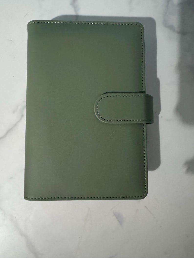 May include: A sage green, rectangular notebook with a rounded flap closure. The cover is smooth and the edges are stitched with a contrasting thread. The notebook is closed with a matching flap and a rounded tab.