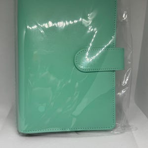 May include: A mint green planner in a clear plastic sleeve. The planner has a smooth, glossy finish and a tab closure. The edges are stitched with a matching thread. The planner is rectangular and appears to be new.