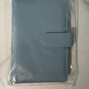 May include: A light blue, rectangular planner with a matching tab closure. The planner is encased in clear plastic. The cover and tab are made of a smooth material, and the edges are stitched.