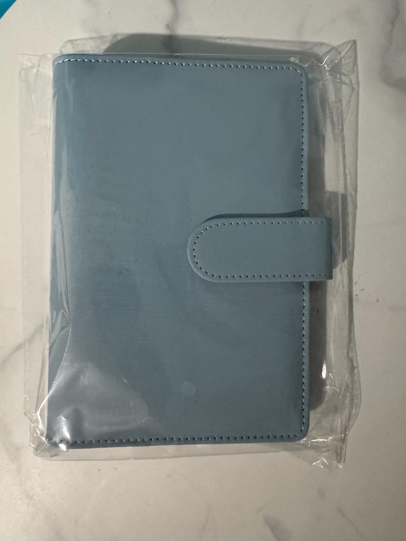 May include: A light blue, rectangular planner with a matching tab closure. The planner is encased in clear plastic. The cover and tab are made of a smooth material, and the edges are stitched.