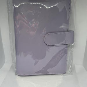 May include: A lavender-colored planner with a glossy finish, encased in clear plastic. The planner features a tab closure on the right side. The cover is smooth and reflects light, suggesting a patent leather or similar material. The overall design is simple and functional.