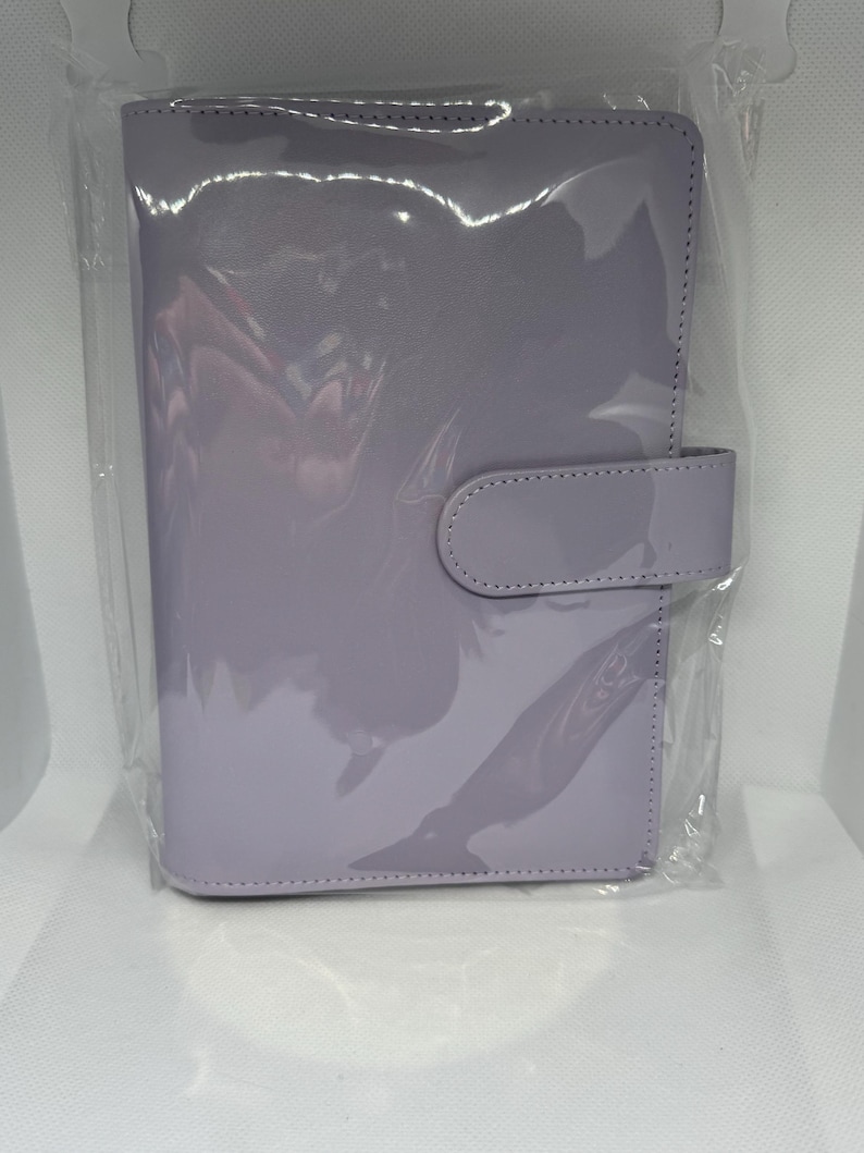 May include: A lavender-colored planner with a glossy finish, encased in clear plastic. The planner features a tab closure on the right side. The cover is smooth and reflects light, suggesting a patent leather or similar material. The overall design is simple and functional.
