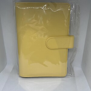 May include: A yellow, glossy, leather-like planner in a clear plastic package. The planner has a rectangular shape with rounded corners and a tab closure. The cover is a solid, bright yellow color.