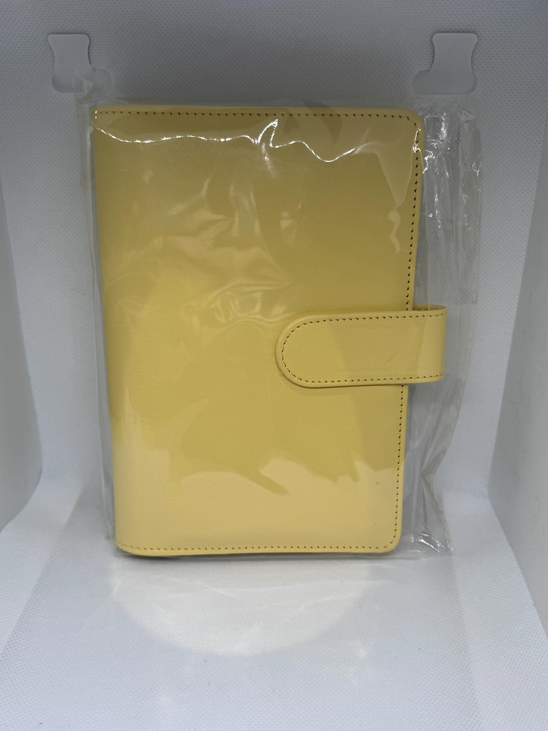 May include: A yellow, glossy, leather-like planner in a clear plastic package. The planner has a rectangular shape with rounded corners and a tab closure. The cover is a solid, bright yellow color.
