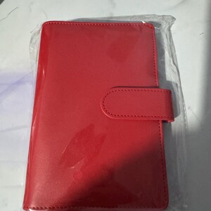 May include: A bright red, rectangular notebook or planner, encased in clear plastic. The cover has a smooth, glossy finish and a rounded flap with a matching snap closure. The item appears new and unused.