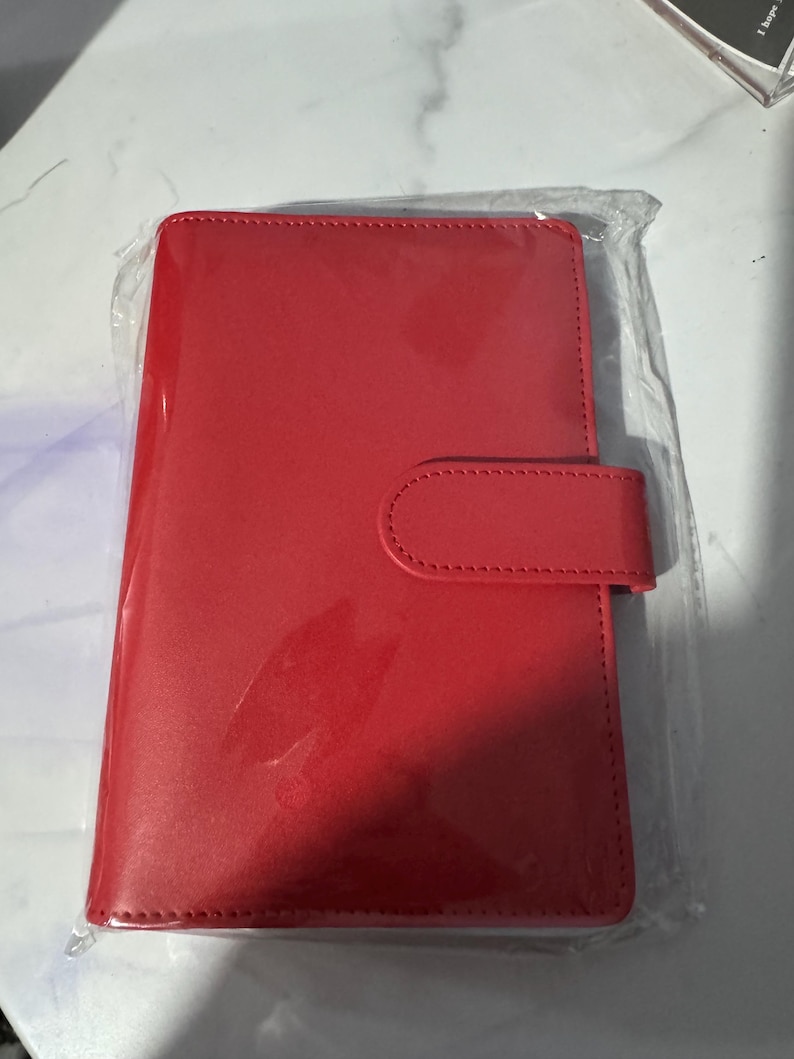 May include: A bright red, rectangular notebook or planner, encased in clear plastic. The cover has a smooth, glossy finish and a rounded flap with a matching snap closure. The item appears new and unused.