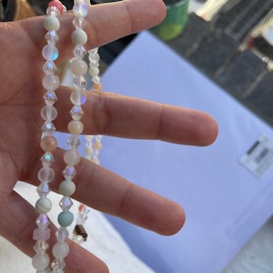 Könnte beinhalten: Zwei schimmernde Perlenketten mit einem Mix aus klaren, weißen, rosa und grünen Perlen. Die Ketten sind übereinander gelegt und werden in einer Hand gehalten.