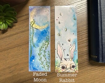 May include: Two watercolour bookmarks. The first bookmark features a crescent moon with stars and green vines on a blue background. The text "Faded Moon" is below the image. The second bookmark features a white rabbit with pink ears and a yellow flower on a blue background with butterflies. The text "Summer Rabbit" is below the image.