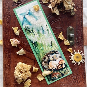 May include: A watercolor bookmark featuring a hedgehog with mushrooms, set against a green and white background. The bookmark is bordered in green and rests on a brown leather journal with floral embossing. Dried flowers and petals surround the bookmark.