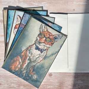 May include: A stack of watercolor art cards featuring a fox wearing glasses and a bow tie. The fox is depicted in shades of orange, brown, and white against a muted blue and green background. A white envelope is next to the cards.