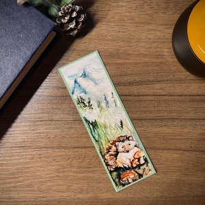 May include: A watercolor bookmark featuring a hedgehog nestled among mushrooms and tall grass. The top of the bookmark depicts a mountain scene with snow-capped peaks. The bookmark has a light green border and is set on a wooden surface.