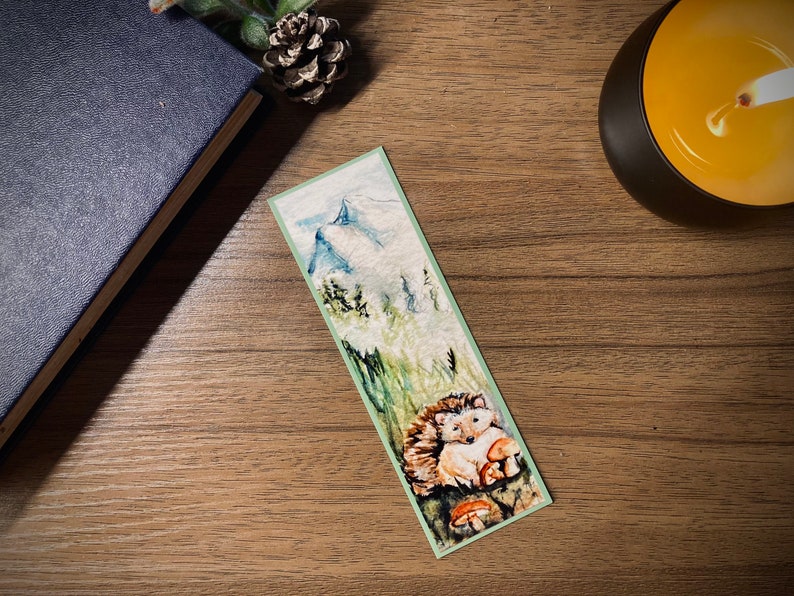 May include: A watercolor bookmark featuring a hedgehog nestled among mushrooms and tall grass. The top of the bookmark depicts a mountain scene with snow-capped peaks. The bookmark has a light green border and is set on a wooden surface.