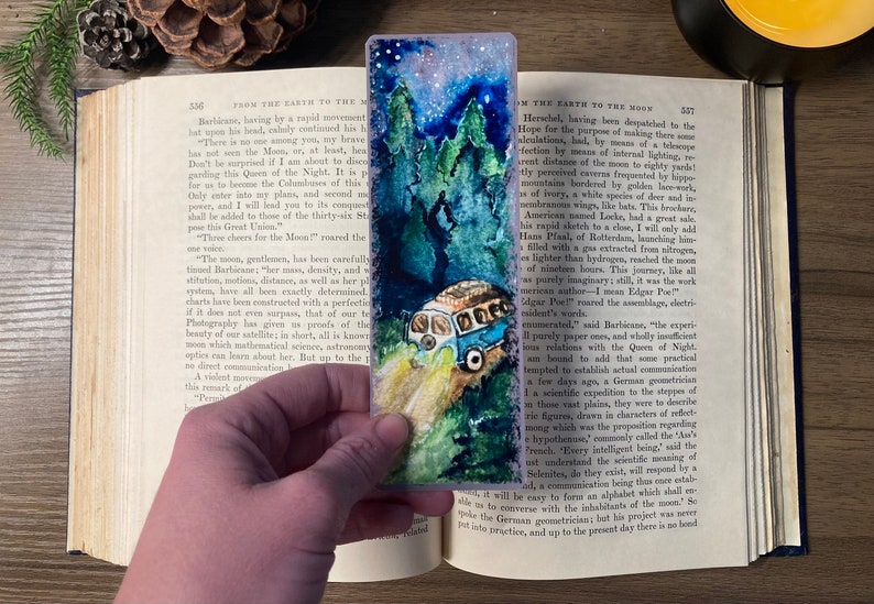 K&ouml;nnte beinhalten: Ein Aquarell-Lesezeichen mit einem alten Van, der nachts durch einen Wald f&auml;hrt. Der Himmel ist mit Sternen gef&uuml;llt und die Scheinwerfer des Vans beleuchten den Weg vor ihm.