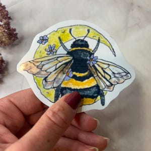 Könnte beinhalten: Ein Aufkleber mit einer Aquarellillustration einer Hummel mit gelbem und schwarzem Körper, vor einem gelben Halbmond mit kleinen lila Blumen. Die Biene hat detaillierte Flügel und Fühler. Der Aufkleber wird von einer Hand gehalten.
