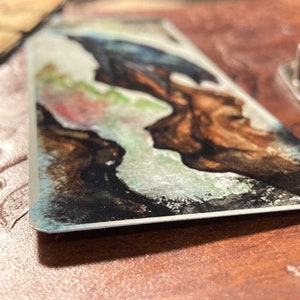 May include: A watercolor painting on a metal card with a blue and green abstract design. The design features a dark brown shape with a white outline.