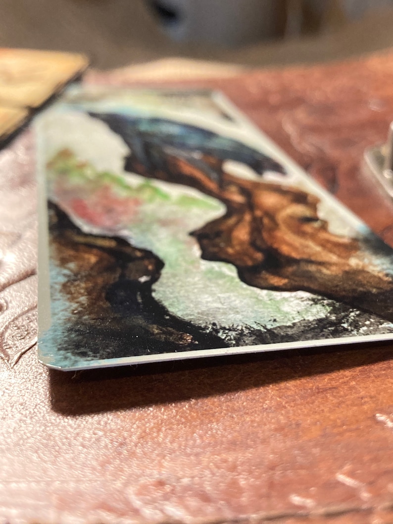 May include: A watercolor painting on a metal card with a blue and green abstract design. The design features a dark brown shape with a white outline.