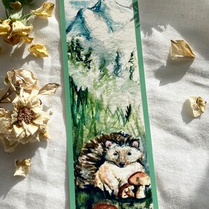 May include: Watercolor bookmark featuring a hedgehog and mushrooms in a forest setting. The top of the bookmark depicts a mountain range with a misty sky. The bookmark has a green border.