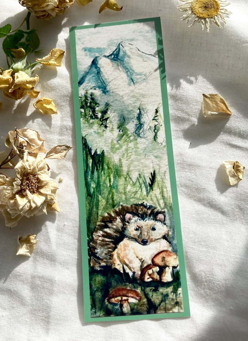 May include: Watercolor bookmark featuring a hedgehog and mushrooms in a forest setting. The top of the bookmark depicts a mountain range with a misty sky. The bookmark has a green border.