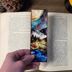 K&ouml;nnte beinhalten: Ein Aquarell-Lesezeichen mit einem sternenklaren Nachthimmel, einem Halbmond, einer Bergkette und einem Zelt mit einem Lagerfeuer. Das Lesezeichen hat einen braunen Holzrahmen.