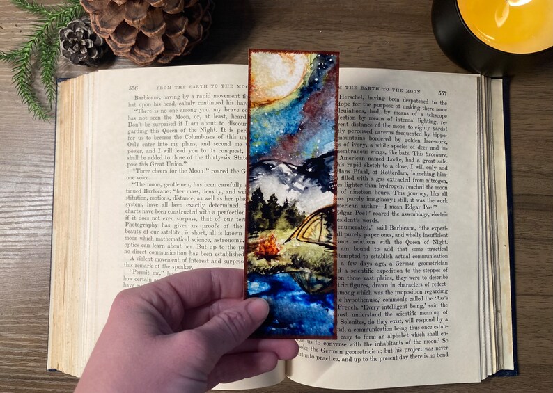 K&ouml;nnte beinhalten: Ein Aquarell-Lesezeichen mit einem sternenklaren Nachthimmel, einem Halbmond, einer Bergkette und einem Zelt mit einem Lagerfeuer. Das Lesezeichen hat einen braunen Holzrahmen.
