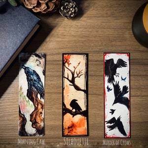 May include: Three watercolor bookmarks featuring black crows. The first bookmark shows a single crow perched on a branch with the text "Morning Caw". The second bookmark shows a crow perched on a branch with the text "Silhouette". The third bookmark shows a group of crows in flight with the text "Murder of Crows".
