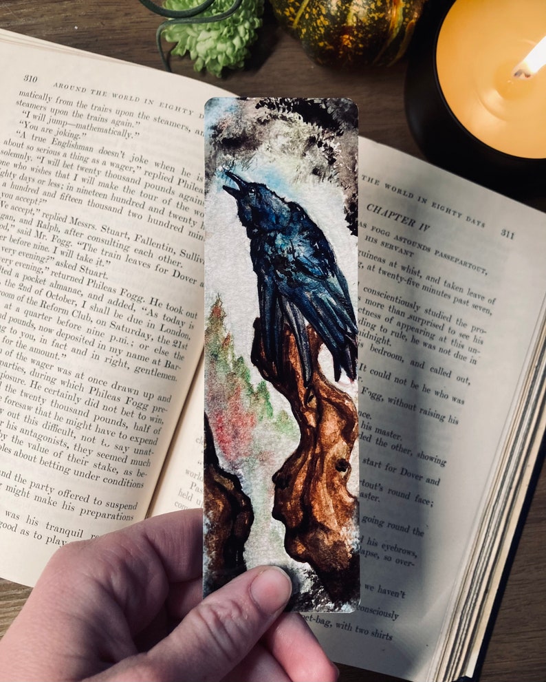 May include: A watercolor bookmark featuring a raven perched on a tree branch. The raven is dark blue with a black beak and eyes. The background is a soft, watercolor wash of blues and greens. The bookmark is titled "Chapter IV" and features the text "As Fogg Astounds Passepartout, His Servant".