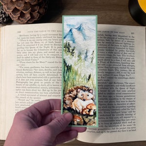 May include: Watercolor bookmark featuring a mountain landscape with a hedgehog and mushrooms. The bookmark is held over an open book with text. The bookmark has a green border.