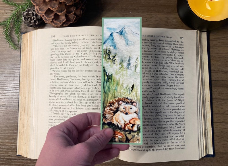 May include: Watercolor bookmark featuring a mountain landscape with a hedgehog and mushrooms. The bookmark is held over an open book with text. The bookmark has a green border.
