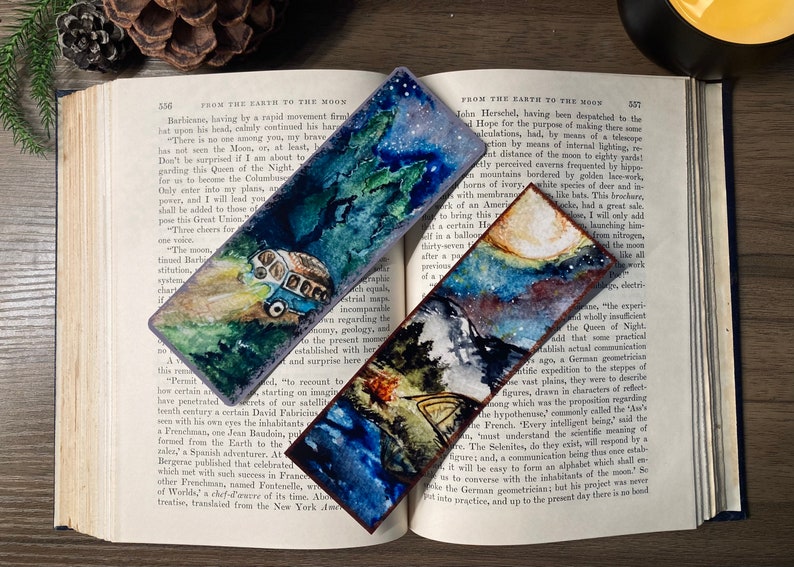 K&ouml;nnte beinhalten: Drei Aquarell-Lesezeichen mit Weltraumthema. Das erste Lesezeichen zeigt einen blauen und gr&uuml;nen Van, der durch einen Wald mit einem sternenklaren Nachthimmel f&auml;hrt. Das zweite Lesezeichen zeigt eine Bergkette mit einem Lagerfeuer und einem sternenklaren Nachthimmel. Das dritte Lesezeichen zeigt eine Bergkette mit einem Vollmond und einem sternenklaren Nachthimmel.