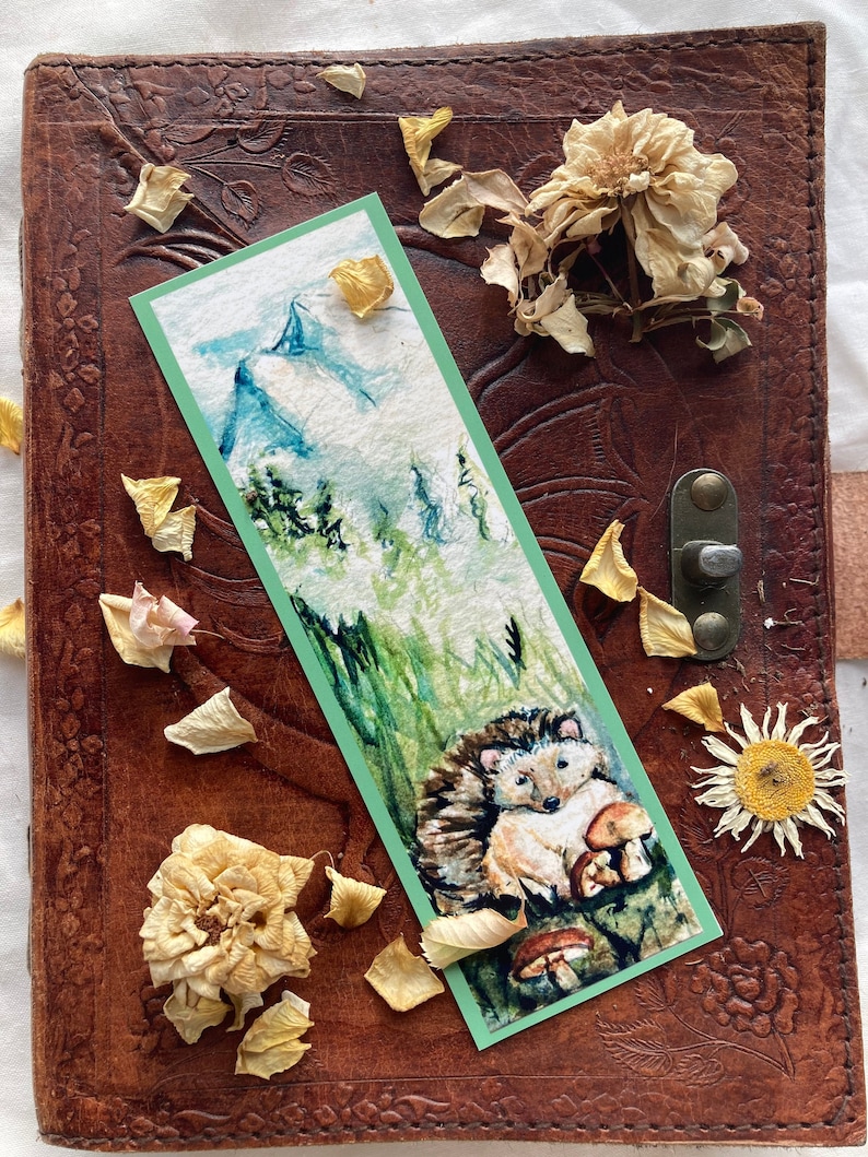 May include: A watercolor bookmark featuring a hedgehog with mushrooms, set against a green and white background. The bookmark is bordered in green and rests on a brown leather journal with floral embossing. Dried flowers and petals surround the bookmark.