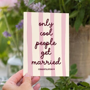 Könnte beinhalten: Eine rechteckige Grußkarte mit cremefarbenem Hintergrund und vertikalen hellrosa Streifen. Der Text in dunkelroter Schreibschrift lautet: only cool people get married. Darunter steht in kleineren Druckbuchstaben: CONGRATULATIONS mit einem kleinen Herz.