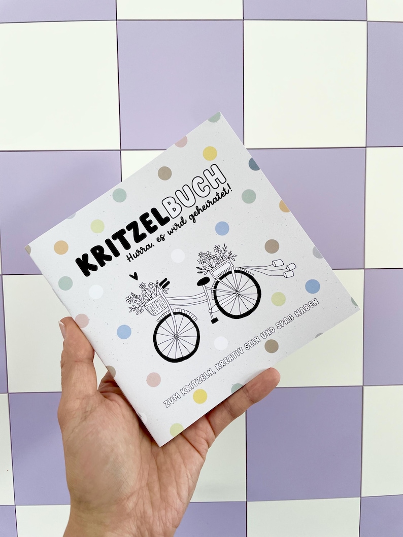 May include: A white booklet with colorful polka dots and the text "KRITZELBUCH" in black. The text "Hurra, es wird geheiratet!" is also in black. The booklet features a black and white illustration of a bicycle with flowers in a basket. The text "ZUM KRITZELN, KREATIV SEIN UND SPA&szlig; HABEN" is in black.